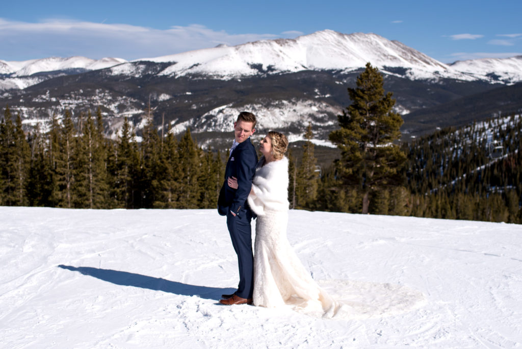 Groom lookin over his shoulder at his bride on the top of the mountain at Breckenridge Ski Resort during their Colorado winter elopement in Breckenridge