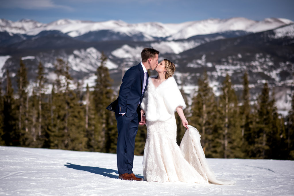 Bride with winter shawl over her wedding dress kissing groom at the top of the mountain in Breckenridge during their ski resort winter elopement in Colorado