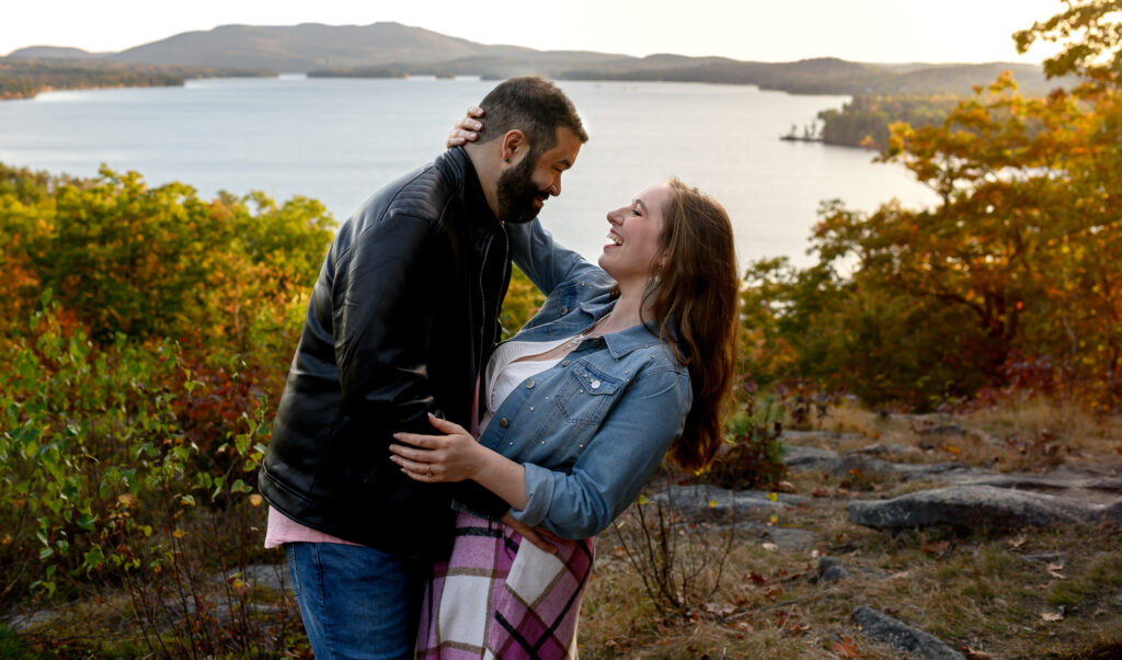 Couple in an embrace and smiling at eachother in front of a lake background in sunapee nh